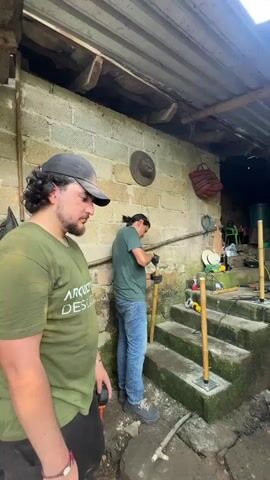 A group of people wearing matching green t-shirts with the words "ARQUITECTS DESCALZOS" are working on building a bamboo railing on a set of concrete stairs leading up to a partially covered outdoor area. Two men are actively working on the railing, one using a power drill and the other using a tool on a bamboo pole. Another person is working inside the covered area. Three other people stand nearby. The ground around the stairs is uneven and partially covered with loose debris and mud. The area is surrounded by trees and vegetation. The sun is shining through the trees.