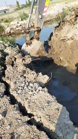 This is a close-up, low-angle shot of a yellow Volvo excavator at work on a sunny day. The excavator's large bucket is digging into the muddy bank of a narrow, shallow body of water, likely a ditch or stream. Dirt and debris are being scooped up and piled onto the bank. The excavator's arm and boom are prominent in the foreground, with the cabin visible further back. The surrounding environment appears to be a construction or excavation site, with loose soil, some rocky debris, and sparse vegetation. In the background, there are hints of distant trees, power lines, and a gravel pile, suggesting a semi-rural or industrial setting. The video is shaky, indicating it was likely recorded handheld.