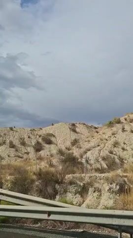 The video shows a view from a vehicle traveling along a road. In the foreground, there is a metal guardrail. Beyond the guardrail, a dry, hilly landscape is visible, characterized by pale, eroded earth and sparse vegetation. The sky above is partly cloudy with a mix of gray and white clouds. The camera pans slightly to the right, revealing more of the hilly terrain.