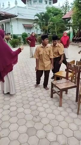 The video shows a group of children, mostly boys in school uniforms, participating in a game of musical chairs outdoors. The ground is paved with hexagonal tiles. Several wooden chairs are arranged in a circle, with one chair missing for each round. The children are shown walking or dancing around the chairs as music plays, and then rushing to sit down when the music stops. Some adults are also present, observing the game. The setting appears to be a schoolyard, with a building in the background. The overall atmosphere is lively and festive.