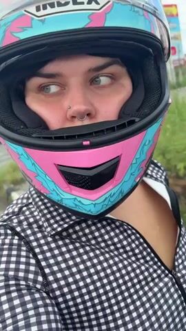 A person wearing a pink and blue patterned full-face motorcycle helmet and a black and white checkered jacket is shown from the chest up. The person has blue-green eyes and is looking at the camera. A nose ring and septum piercing are visible. The video then cuts to a first-person view from behind the helmet, showing a street scene with cars and motorcycles in traffic under a cloudy sky. The video then returns to the close-up of the person's face.