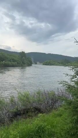 A wide, choppy river flows from left to right. Dense green trees line the banks of the river. In the distance, across the river, several houses are nestled amongst the trees at the base of a large, forested hill. Smoke rises from one of the houses, indicating a fire or chimney. The sky is overcast with gray clouds. In the foreground, tall green grass and brown reeds grow along the riverbank.