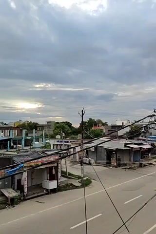 The video shows a street scene with several small shops lining one side of the road. The sky is overcast with scattered sunlight peeking through the clouds. The shops appear to be closed, with their shutters down. There are electrical wires running overhead and a few utility poles. A person is visible near one of the shops, standing next to a green trash bin. The road is empty of vehicles. The buildings are simple structures, typical of a commercial area in a town or city. The overall impression is a quiet, possibly early morning or late afternoon scene.