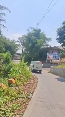 The video begins with a view from a moving vehicle on a paved road. Lush green vegetation lines the left side of the road, with a white van in front of the camera also traveling on the road. To the right, there are some buildings and more greenery. As the video progresses, the van continues to drive away from the camera. A traffic signal, which is red, appears on the left side of the road. Further down the road, a red convex mirror is attached to a pole on the left. The sky is clear and blue. The overall environment appears to be a rural or suburban area with a lot of trees and vegetation.