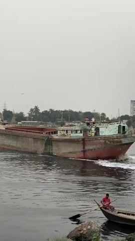 A large, long cargo ship, possibly a barge, moves from right to left across a wide river under an overcast sky. The ship has a red hull with an aged, weathered appearance, and its upper deck structure is painted with faded green and blue. White foam trails behind its stern as it moves. Several people are visible on the deck of the large vessel.

In the foreground, closer to the viewer, a small, dark wooden rowboat is positioned, facing right. A man wearing a red shirt is seated in the rowboat, holding an oar, seemingly observing the larger ship. The river water appears dark and slightly murky.

The distant riverbank features lush green trees and vegetation, with some buildings and what appear to be power lines or utility poles visible through the haze. A bird can be seen flying in the upper left portion of the sky. The scene depicts typical river activity in Tarabo, Bangladesh, during what appears to be daytime, under cloudy conditions. Faint, unreadable text and numbers are visible on the side cabin structure of the large ship.