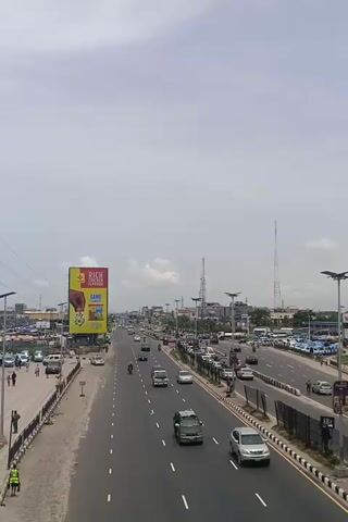The video shows a busy highway in a city on a sunny day. Traffic moves along the multiple lanes.  Buildings and billboards line the side of the road.  The camera is positioned high, giving an elevated view of the scene.  The sky is overcast, creating a slightly hazy atmosphere.