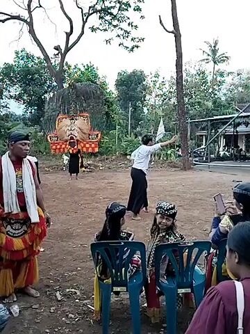 The video shows a cultural performance taking place outdoors in a rural or semi-rural environment.  A large, decorative mask is being carried by a person wearing black clothing. A man in traditional attire, including a white scarf and colorful patterned pants, stands to the side, seemingly observing the performance. A woman in a white shirt and dark pants appears to be directing the performance with arm gestures.  Several people are seated in plastic chairs, some of whom are recording the performance on their phones. The setting includes a dirt ground, trees, and some simple buildings in the background.