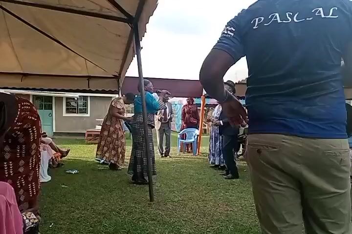 A group of people are gathered outdoors under a canopy. In the center, a woman in a blue top and patterned skirt is speaking into a microphone, gesturing with her free hand. To her right, a man in a suit and hat holds a microphone and appears to be listening or speaking. To the left of the central woman, another woman in a patterned dress is also present, and further left, a woman in a red and white patterned dress is visible. In the foreground, the back of a man wearing a blue shirt with "PASCAL" printed on it is facing away from the camera. The ground is covered in grass, and several chairs are arranged in the background. A building with windows is visible behind the gathering. The sky is overcast.