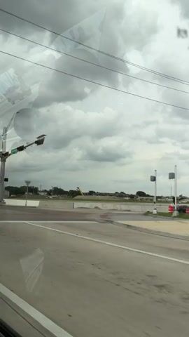 The video shows a view from a vehicle driving on a bridge with an overcast sky. On the left side of the road, there is a traffic light and street signs. The traffic light has green and yellow signals. The street signs indicate "Lyndon B. Johnson" and "Skillman". There is also a street sign with a red circle and a diagonal line, indicating no U-turns. Further down the road, a yellow construction crane is visible. The bridge has a modern, white arched structure with diagonal supports. There are also white concrete barriers along the sides of the bridge. The road is paved and has lane markings. A billboard can be seen in the distance.