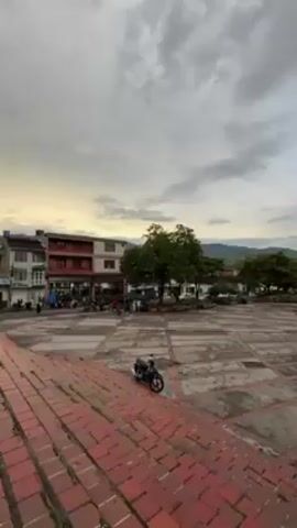 The video shows a wide shot of a large, brick-paved square in a town. In the foreground, there are brick stairs leading down to the main level of the square. A black motorcycle is parked on the edge of the stairs.

In the center of the square, there is a large tree with bare branches, taking up a significant portion of the view. People can be seen walking around the square, though they appear small due to the distance.

Buildings and smaller trees surround the perimeter of the square. In the background, rolling green hills are visible under a cloudy sky. The sky is bright, suggesting it might be early morning or late afternoon.