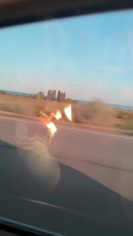 The video shows a blurry, fast-moving view from inside a vehicle. Through the window, a road with a white line is visible, running parallel to the direction of travel. Beyond the road, a dry, grassy field stretches towards a body of water in the distance. The sky is a clear blue with a few wispy clouds. Sunlight creates bright, distorted reflections on the window, obscuring some of the details of the landscape. The overall impression is one of a journey through a rural or coastal area during the day.