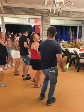 A group of people are dancing in a line, holding hands, and moving from left to right. They are in an indoor space with tables and chairs. The lighting is dim. There is a Turkish flag visible on the wall in the background.