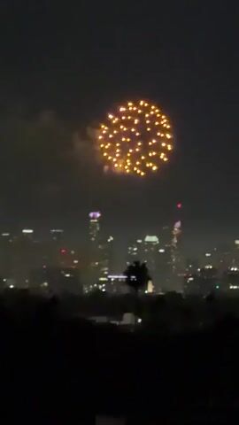 A nighttime aerial view shows fireworks illuminating a city skyline. The fireworks display features bursts of yellow, orange, and red lights against the dark sky. Buildings with illuminated windows are visible in the background. The foreground is dominated by dark silhouettes of trees and possibly hills. The video captures the visual spectacle of the fireworks as they explode in the sky over the urban landscape.