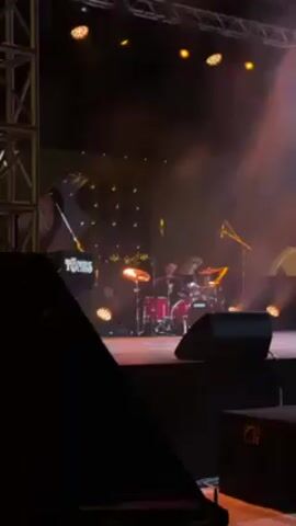 The video shows a drummer performing on stage. The camera zooms in on him. He is wearing a black suit, white shirt, and orange bow tie. He has short blond hair and is smiling slightly. His drum kit is red. The stage is lit with orange lights, and there is smoke in the air. There is also stage equipment and the band name "The Tucos" visible in the background.