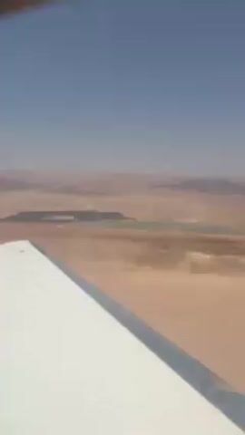 Here is a concise description of the video:

The video shows an aerial view from a small airplane, with a portion of the white wing visible in the foreground. The scenery below appears to be a desert landscape under a clear blue sky. In the distance, there is a rectangular area of green vegetation contrasting sharply with the surrounding arid terrain. The overall atmosphere is calm and sunny, suggesting a flight over a relatively remote area.