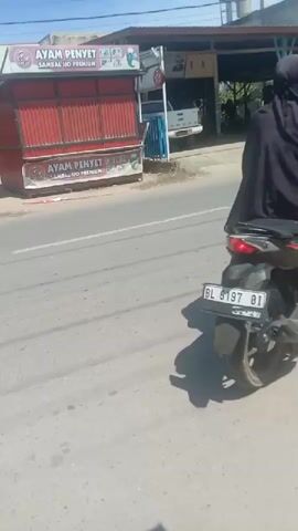A person wearing a black hijab and a patterned garment rides a black motorcycle on a paved road. The motorcycle has a license plate with the characters "BL 8197 BI". The road is bordered by a sidewalk on the right and a row of businesses and buildings on the left. The businesses appear to be open, with some people visible inside. The sky is clear and blue, indicating sunny weather. The overall scene suggests a typical day in a commercial or residential area.