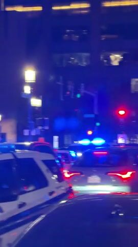 The video shows a line of Boston police vehicles at a stoplight at night. The police cars have their emergency lights flashing. The scene appears to be in a downtown area, with large office buildings visible in the background. One of the police vehicles is a white SUV with blue stripes.  A silver Honda CRV is also visible in front of a police vehicle.