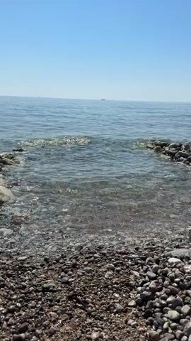 The video shows a view of a calm sea from a pebble beach. The sky is clear and blue. The water is clear and reflects the sunlight. Small waves are gently lapping at the shore. There are some stones and rocks visible on the beach. In the distance, a small boat or ship can be seen on the horizon. The overall scene is peaceful and serene.