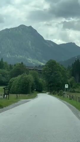 The video shows a winding paved road leading through a lush green landscape with mountains in the background. The road is bordered by wooden fences and grassy fields, with numerous trees and dense forestation on either side. In the distance, a large bridge or viaduct is visible spanning across a valley, and a mountain peak dominates the horizon, partially obscured by dramatic, cloudy skies. There is a bus stop sign with the letter "H" on it to the right of the road, and another road sign with a "20" inside a circle indicating a speed limit. The overall impression is one of a scenic drive through a rural, mountainous region, likely in the Alps given the location context. The weather appears overcast, suggesting it might be morning or late afternoon on a cloudy day. There are no people visible in the footage. The atmosphere is peaceful and tranquil.