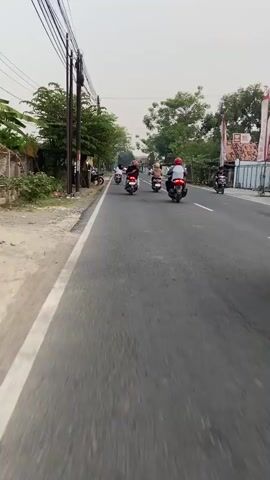 This media file captures a group of people riding motorcycles on a road in Wirosari, Indonesia. The subjects are primarily individuals on scooters and motorcycles, many of whom are wearing helmets. The scene is a paved road with a white line marking the lane. On the left side of the road, there are trees and some simple structures, possibly stalls or small buildings. On the right side, more trees and houses are visible. The activity is clearly travel on motorcycles, with several vehicles in motion.

Notable details include the time of day, which appears to be late afternoon or early evening, given the soft lighting and overcast sky. The weather seems to be calm, with no indication of rain. The location context of Wirosari, Indonesia, is provided. The overall mood appears to be casual and everyday, with people going about their business. There is no visible text within the media file. The individuals are dressed in casual clothing, and some have bags or items on their motorcycles. The road surface shows some wear and tear, with a few potholes visible. The presence of a pedestrian crossing adds a common urban/suburban element to the scene.