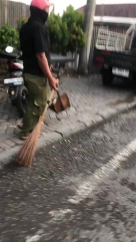 A person wearing a pink hat, black shirt, and green pants is sweeping the ground with a broom and dustpan. They are standing next to a parked motorcycle and a vehicle with a cargo bed. The ground appears to be wet and there is a paved pathway. Trees and bushes are visible in the background.
