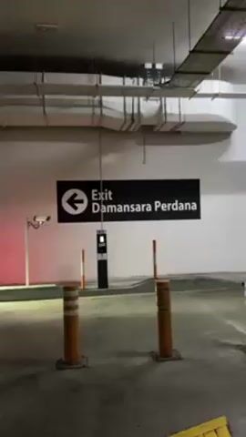 The video shows the interior of a parking garage. Initially, the camera faces a sign reading "Exit Damansara Perdana" with an arrow pointing left. The area is lit by overhead fluorescent lights, and the garage has a concrete floor with multiple expansion joints. There's a parking gate arm and an access control kiosk nearby. The camera then slowly pans to the left, revealing a ramp leading to the outside. Yellow arrows are painted on the ramp to guide traffic. Outside, a road with parked cars can be seen.