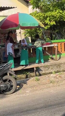 A street-level outdoor scene shows a small roadside stall with a multicolored umbrella providing shade. Three children in green pleated skirts and white headscarves are standing near the stall. One child, in the center, is interacting with a woman behind the stall. Another child stands to the left, also near the stall. A third child, wearing a patterned shirt and green pants, is to the right. A motorcycle is partially visible in the foreground on the left. The environment appears to be a sunny day with greenery and a building in the background.
