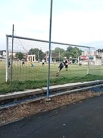 The video shows a soccer field with players during what appears to be a game. There are a few players near one goal, with one on the ground. The camera pans slightly to show a nearby road and a building in the background, then pans back to show the field from a different angle, revealing more players. The field is green and the sky is overcast.