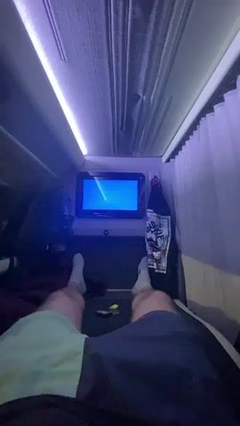 The video shows a first-person perspective of a person lying down on a bed, facing a small screen. The person is wearing shorts and socks. The screen displays a blue background with some yellow and white graphics, possibly from a video or game. The environment appears to be inside a vehicle, possibly a bus or a campervan, with overhead lighting that is illuminated with a purple hue. There are curtains visible on the sides and top, suggesting it is nighttime or the curtains are drawn for privacy. A backpack is visible on the left side of the frame. The overall impression is of someone relaxing and watching something in a confined space.