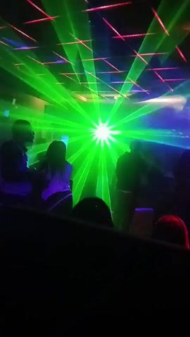 The video shows a group of people at what appears to be a club or concert venue. The scene is dimly lit with colorful lighting including green, red, and blue lasers. The people are in silhouette, making it difficult to discern specific details about them. The video is filmed from a low angle, potentially from a seated position. The setting suggests a lively atmosphere, possibly with music and dancing.