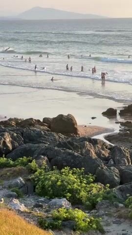 The video shows a beach scene with people swimming and surfing in the ocean. In the foreground, there are rocks and vegetation. The sun appears to be setting, casting a warm glow on the scene. There are several people visible on the beach, including a group of children playing in the sand and a man sitting with a woman. A dog is also present on the beach, interacting with the people. The water is choppy with small waves, and there are several surfers in the water. In the distance, a mountain range is visible against the horizon. The overall atmosphere is peaceful and relaxed.