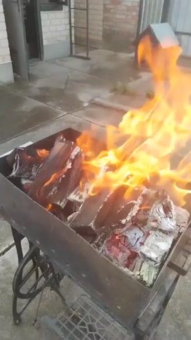 A shot of a metal barbecue grill filled with burning wood. The flames are orange and yellow and rise from the wood. The grill is on a metal stand. The scene is outdoors with a paved surface in the foreground. In the background, there is a brick building and a small dog house. The lighting suggests it is daytime.