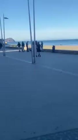 The video shows a seaside promenade on a sunny day. There are people walking along the paved path, with palm trees lining the side. Cars are visible on the road next to the promenade. Street lights are spaced along the path. The video ends with a view of the beach and the ocean.