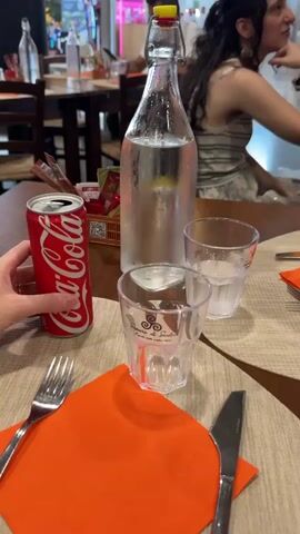 A close-up shot shows a person's hand holding a red Coca-Cola can and pouring the dark brown liquid into a clear plastic cup. The cup is partially filled with the cola and has some foam on top. In the background, a large glass bottle of water with a slice of lemon is visible, along with another glass filled with water. The scene appears to be set at a table in a restaurant, with an orange napkin and cutlery laid out. Another person is partially visible in the background, seated at a table.