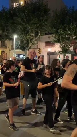 The video shows a street parade at dusk in Arenys de Mar, Spain. A group of people, including adults and children, are marching and playing percussion instruments. Most of them are wearing black t-shirts with a colorful logo featuring the word "RASS" and some stylized green leaves. The participants are playing a variety of instruments, including drums, tambourines, and what appears to be a small cymbal. The atmosphere seems lively and festive, with the participants focused on their music and the overall rhythm of the parade. The street is lined with trees and buildings, suggesting a typical urban or town setting. Streetlights and building lights are illuminated, indicating it is nighttime or late evening. The sky is a dark blue, confirming the time of day. The people are walking on a paved road, and there are some traffic cones and a pedestrian crossing visible in the background, further grounding the scene in a public street environment.