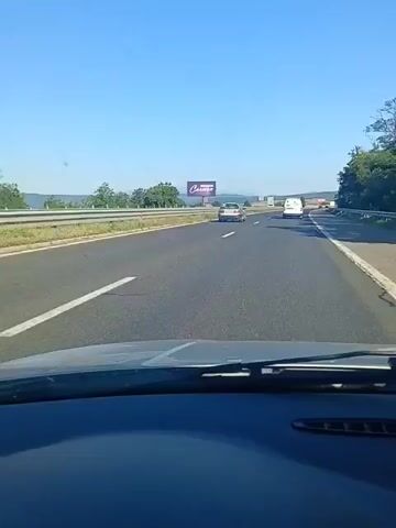 The video begins from the perspective of someone inside a car, showing the dashboard and windshield. The car is on a highway with multiple lanes. In the distance, there is a billboard with the word "Carmen" and a smaller word above it that is difficult to read. Several cars are visible on the highway, including a black SUV, a white car, and other vehicles further ahead. The weather appears to be clear with a bright blue sky. Trees and vegetation line the side of the highway, and hills or mountains are visible in the far distance. The video shows a smooth driving experience on a well-maintained road.