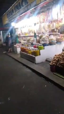 The video captures a street scene, likely in Cisarua, Indonesia, at night. The primary focus is a brightly lit storefront that appears to be a small shop or stall selling various goods. The shop is packed with products displayed on shelves and in bins. We see rows of bottled items, possibly food items like pickles or preserves, in shades of orange, yellow, and green. Above these, there are numerous packets of snacks or dried goods hanging. The shop is well-lit by fluorescent lights, creating a warm and inviting atmosphere, and a prominent yellow sign with text is visible above the merchandise.

In front of the shop, a concrete pathway or sidewalk is visible, and further forward, a dark asphalt road. Two individuals are seen standing near the entrance of the shop. One appears to be wearing a dark shirt and pants, while the other is in a blue t-shirt and shorts. They seem to be interacting or browsing.

To the left of the main shop, another smaller stall or display is visible, featuring a sign that says "CHOCO LAVA". This suggests a variety of offerings beyond just the items in the main shop. There is also what looks like a freezer unit visible, possibly for cold drinks or ice cream.

The overall impression is a bustling night market or a roadside shop, common in many Indonesian towns. The abundance of goods and the lighting suggest a lively commercial area. The time of day is clearly night due to the dark surroundings and the artificial lighting being the primary illumination. The presence of an outdoor stall and the general ambiance points towards a warm climate, typical of Indonesia.