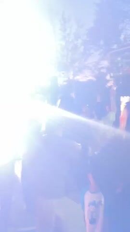 The video shows a gathering of people outdoors at dusk. The sky is overcast, and trees are visible in the background, along with some lights. Several individuals are dancing and raising their arms in the air, suggesting a celebratory atmosphere. There appears to be a stage or performance area with lighting equipment and speakers. Some people are holding phones, possibly recording the event. The ground is a dark surface, possibly a court or paved area. There are also some tents or shelters set up. The overall impression is of a lively outdoor event.