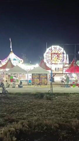 The video shows a nighttime scene at what appears to be a fair or festival. There are several tents set up, some with lights and banners. In the background, a large, illuminated Ferris wheel is visible, along with people gathered around it. A motorcycle is parked in the foreground on the left. The overall atmosphere is festive and bustling, with lights and activity suggesting a public event.