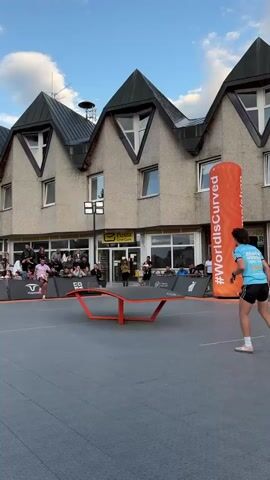 A person in a blue shirt and black shorts is playing table tennis outdoors on a grey surface. They are in the process of hitting the ball over the orange, curved table. In the background, there is a building with a grey facade and dark triangular roofs. A large orange inflatable tube with white text that says "#WorldisCurved" is standing to the right of the table. A crowd of people is watching the game from behind a barrier in front of the building. The sky is blue with some clouds.