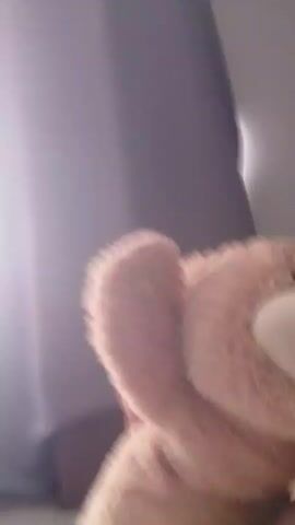 A close-up, low-angle, handheld shot focuses on a large, fluffy, light brown teddy bear. The bear's left paw is raised, and its head is turned to the right, revealing a smiling face with a dark brown nose and stitched eyes. The bear appears to be held by an unseen person, with a glimpse of skin visible beneath its body.

The background consists of a dark gray curtain on the left and a lighter gray wall on the right, suggesting an indoor setting. The lighting is dim, creating shadows and a somewhat intimate atmosphere. No specific time of day or weather conditions are discernible. There are no visible text elements in the media file. The overall impression is of a casual, possibly private moment, with the teddy bear as the central, anthropomorphized subject.