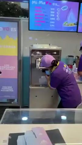 A woman in a purple uniform and cap works behind the counter of an ice cream or frozen yogurt shop. She is using a machine to dispense a frozen dessert into a blue cup. The sign on the machine and the wall in the background advertise the product, mentioning "toppings" and prices for different sizes. The woman takes the cup after filling it. The shop is clean and well-lit.