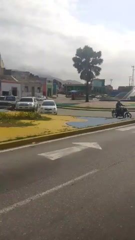 The video shows a roundabout with a fountain in the center of a town square, featuring a statue. A person on a motorcycle is seen on the periphery of the roundabout. The roundabout is painted with various colors, and the buildings surrounding the square are a mix of colors and architectural styles. There are hills in the background under an overcast sky. The video is taken from a slightly low angle, showing a pedestrian crosswalk in the foreground.