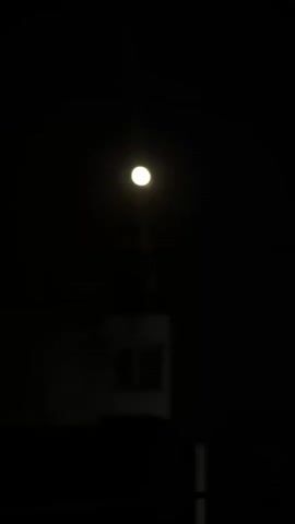 This video clip captures a nighttime scene illuminated by a bright, full moon. The moon is the dominant subject, appearing as a luminous orb against a completely dark sky. While the moon itself is clear, the surrounding environment is indistinct due to the darkness.

Beneath the moon, faint outlines of structures are visible. These appear to be buildings with some form of windows or openings. The overall impression is of an outdoor setting, possibly a residential or semi-urban area. The low visibility and the presence of artificial light sources (implied by the moon's brightness, which could be enhanced by atmospheric conditions or simply the clear night) suggest that the photograph was taken at night.

There are no people or specific objects clearly identifiable in the scene. The focus is entirely on the celestial body and the obscured landscape below. The mood is quiet and serene, characterized by the stark contrast between the brilliant moon and the deep darkness. The location context places this scene in Paralimni, Cyprus, suggesting a Mediterranean night sky. No text is visible in the clip.