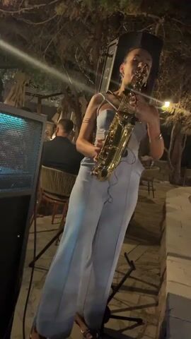 A woman plays a saxophone outdoors at night. She is wearing a light blue jumpsuit and silver sandals. She is standing in front of a large speaker with blue lights. There are trees and other people in the background.