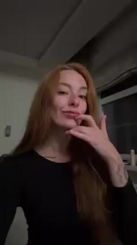 The video shows a woman with long, red hair and fair skin. She is wearing a black, long-sleeved shirt. She touches her lips with her index finger and then looks directly at the camera with a slight smile. The background includes a window with curtains, a partial view of a mirror, and what appears to be a hotel room. The lighting is somewhat dim. There is music playing in the background.