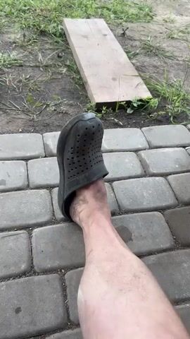 The video shows a person's leg and foot wearing a dark gray Croc shoe. The foot is resting on a paved surface made of rectangular gray stones. In the background, there is dirt, grass, and a wooden plank lying on the ground. The video is shot from a first-person perspective.