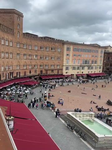The video shows a large, open piazza in an old European city, possibly in Italy. The piazza is paved with light-colored stone in a pattern of lines. Several buildings surround the piazza. Some buildings are light-colored brick with many windows. One building is a large brick structure with a tall clock tower. There are many people in the piazza, mostly walking around. Some people are sitting or lying on the ground. There is a fountain with a green pool of water. Red awnings are visible along the edges of the buildings. Some clouds are visible in the sky.
