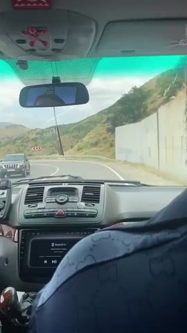 A video recorded from the passenger seat of a car shows the road ahead and the driver's hands and the dashboard. The car is moving along a road that curves to the left, with green hills and mountains on either side. A white retaining wall is visible on the right side of the road. The driver, a man with a beard, is visible from the back. His hands are on the steering wheel. The dashboard of the car is visible, with a navigation system displaying "Bluetooth 5.1". Two air fresheners are hanging from the rearview mirror. The sky is cloudy.