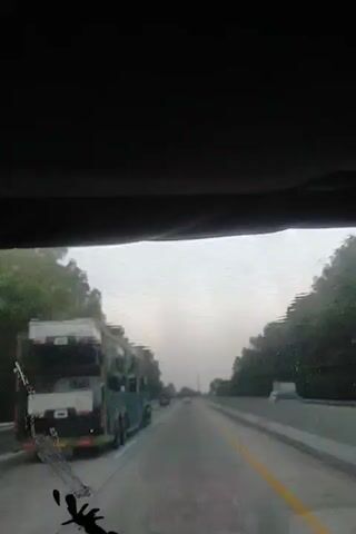 A first-person view from inside a vehicle shows a highway with a large truck on the left side. The sky is overcast, and the windshield is wet, suggesting rain or moisture. Trees line both sides of the highway. The truck appears to be green with a white cargo cover. The road has yellow and white lane markers. The overall impression is of traveling on a highway in potentially inclement weather.
